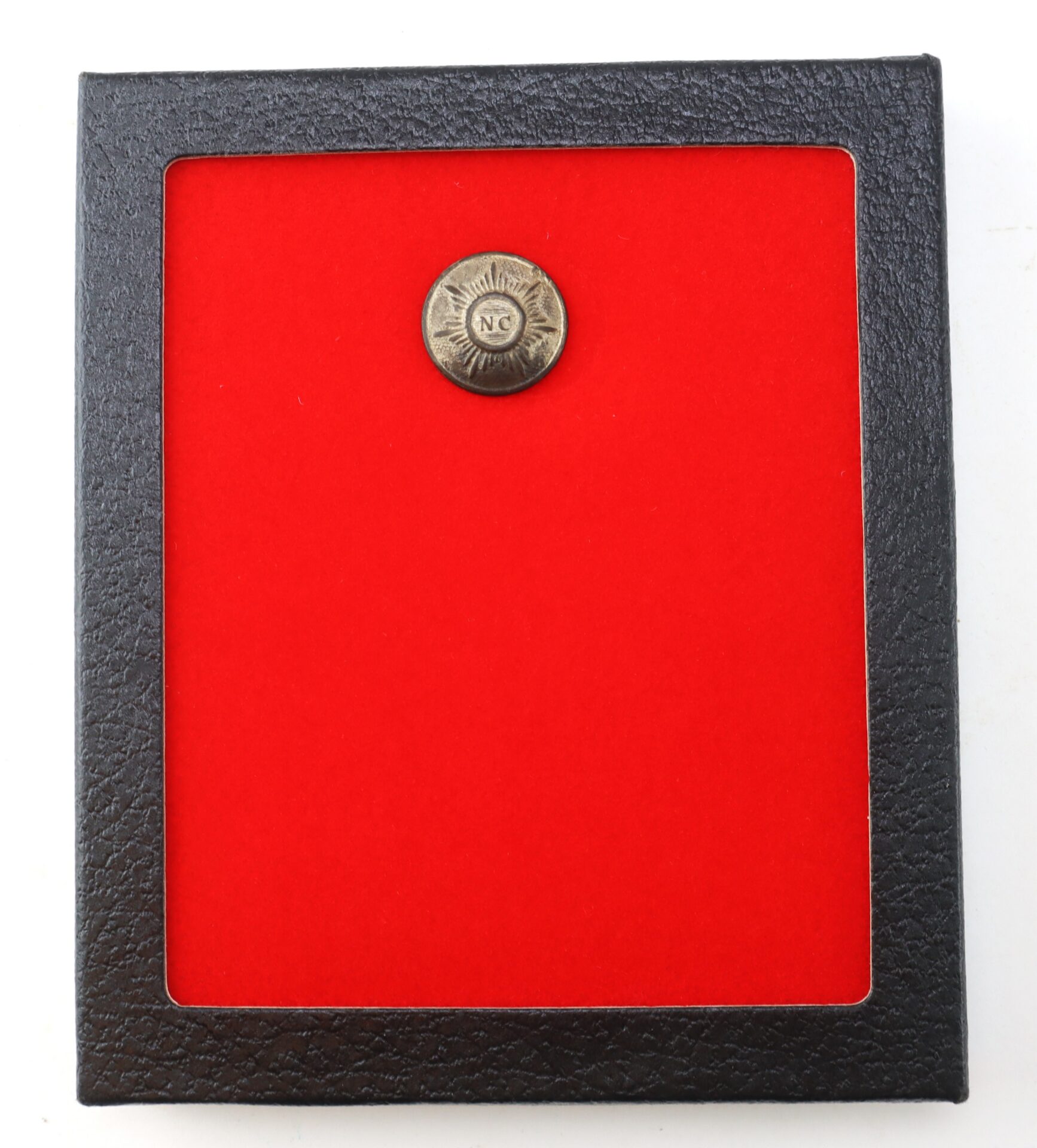 A small round metal button with “NC” on it is displayed in the center of a red background, surrounded by a black rectangular frame.
