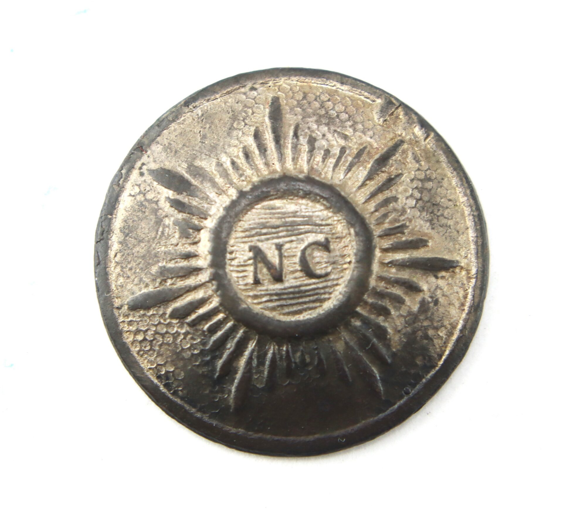Round metal button with a sunburst design and the letters “NC” in the center. The surface appears worn and textured.