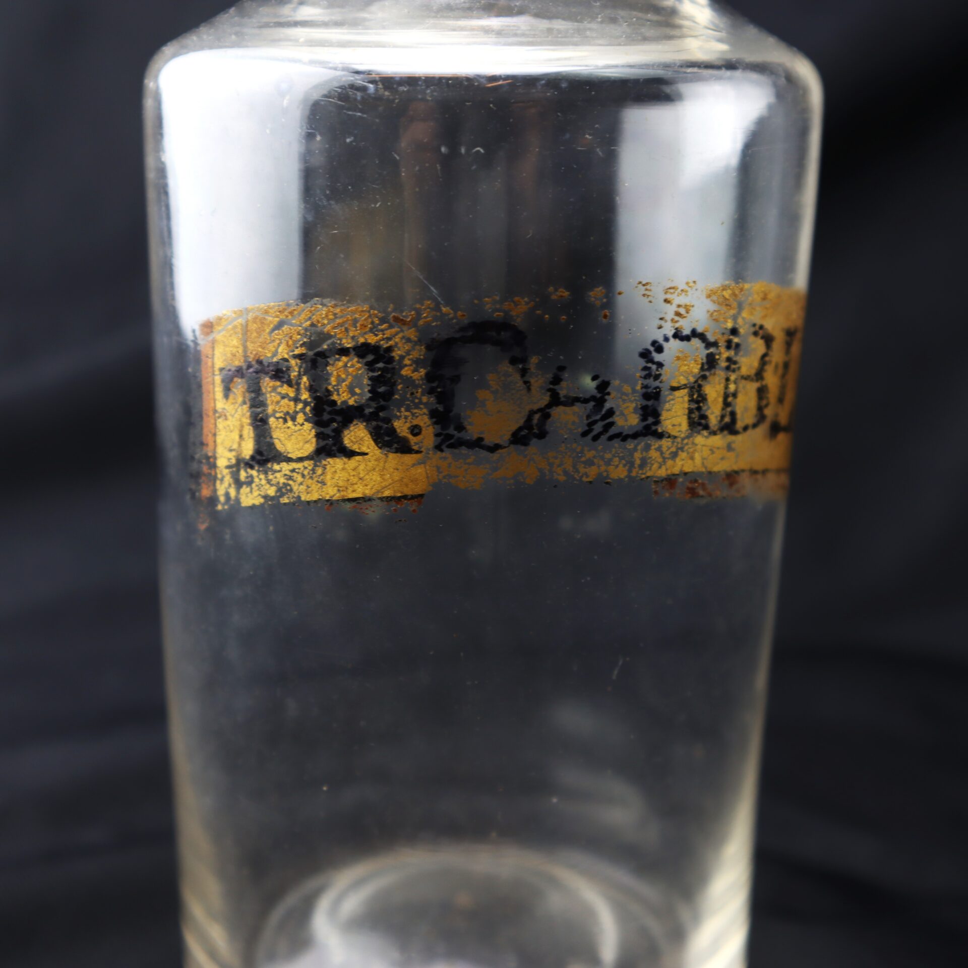 Clear glass bottle with partially faded black and gold label featuring unreadable text, set against a dark background.