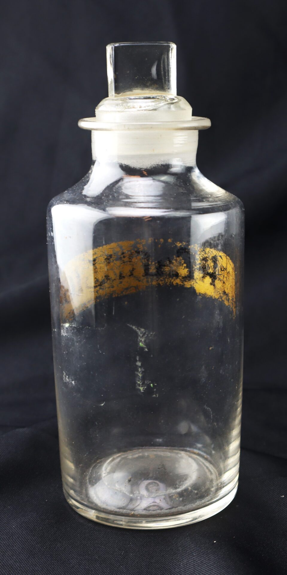 Clear glass bottle with a ground glass stopper and a faded yellow label, set against a black background.