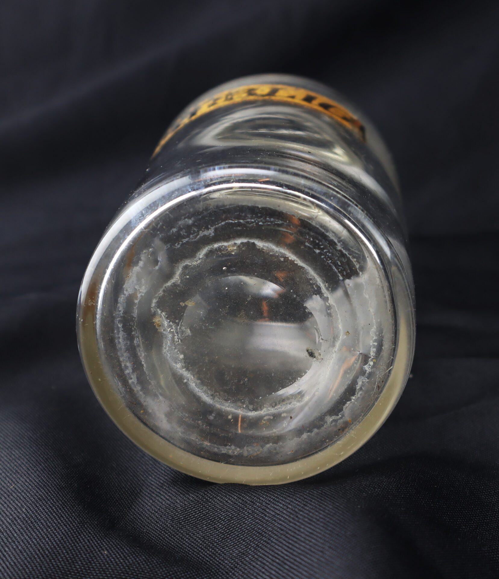 Clear glass jar viewed from the bottom, showing some residue and signs of wear, placed on a black fabric background.