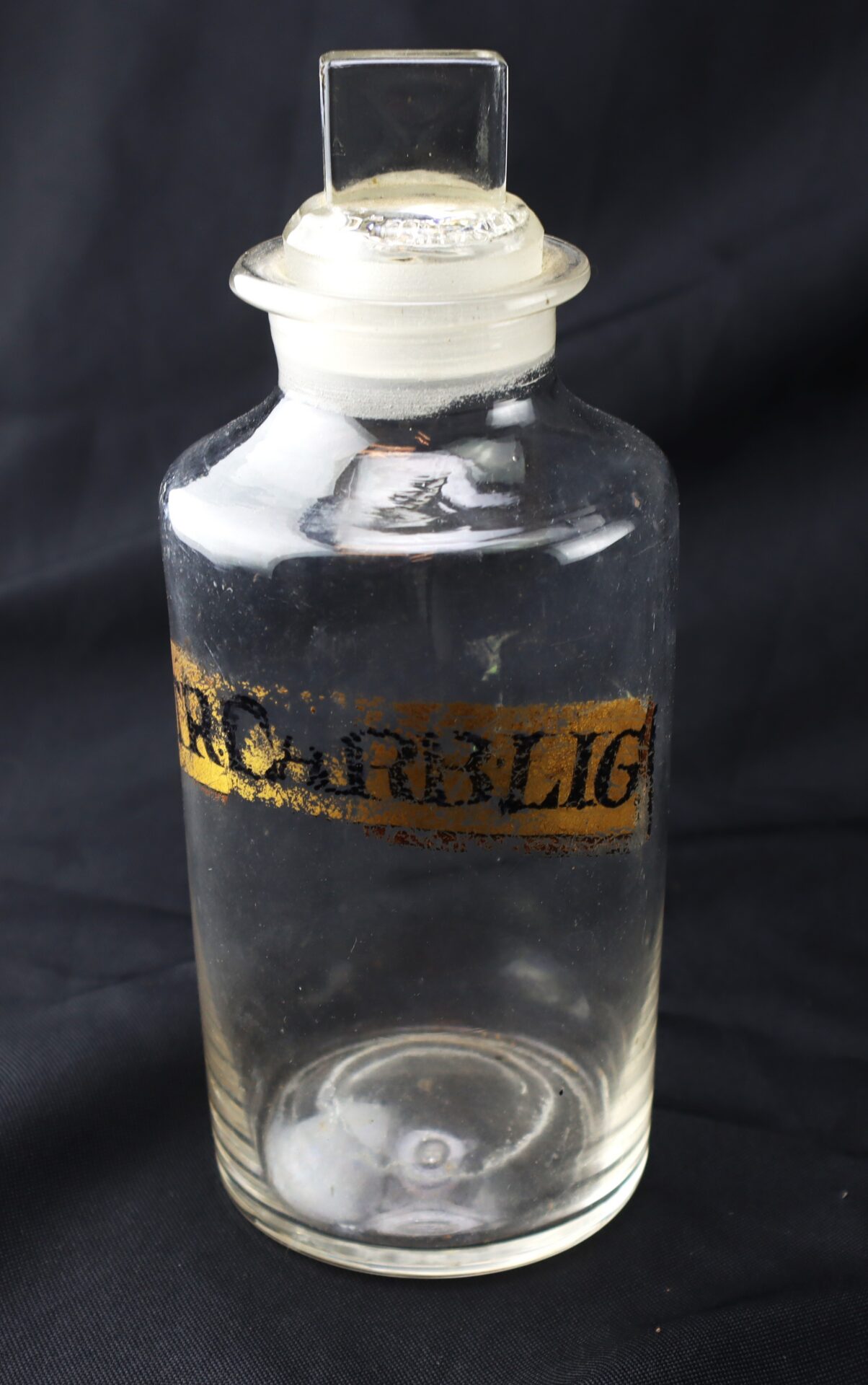 Clear glass apothecary jar with a square stopper and a worn gold and black label on a black background.