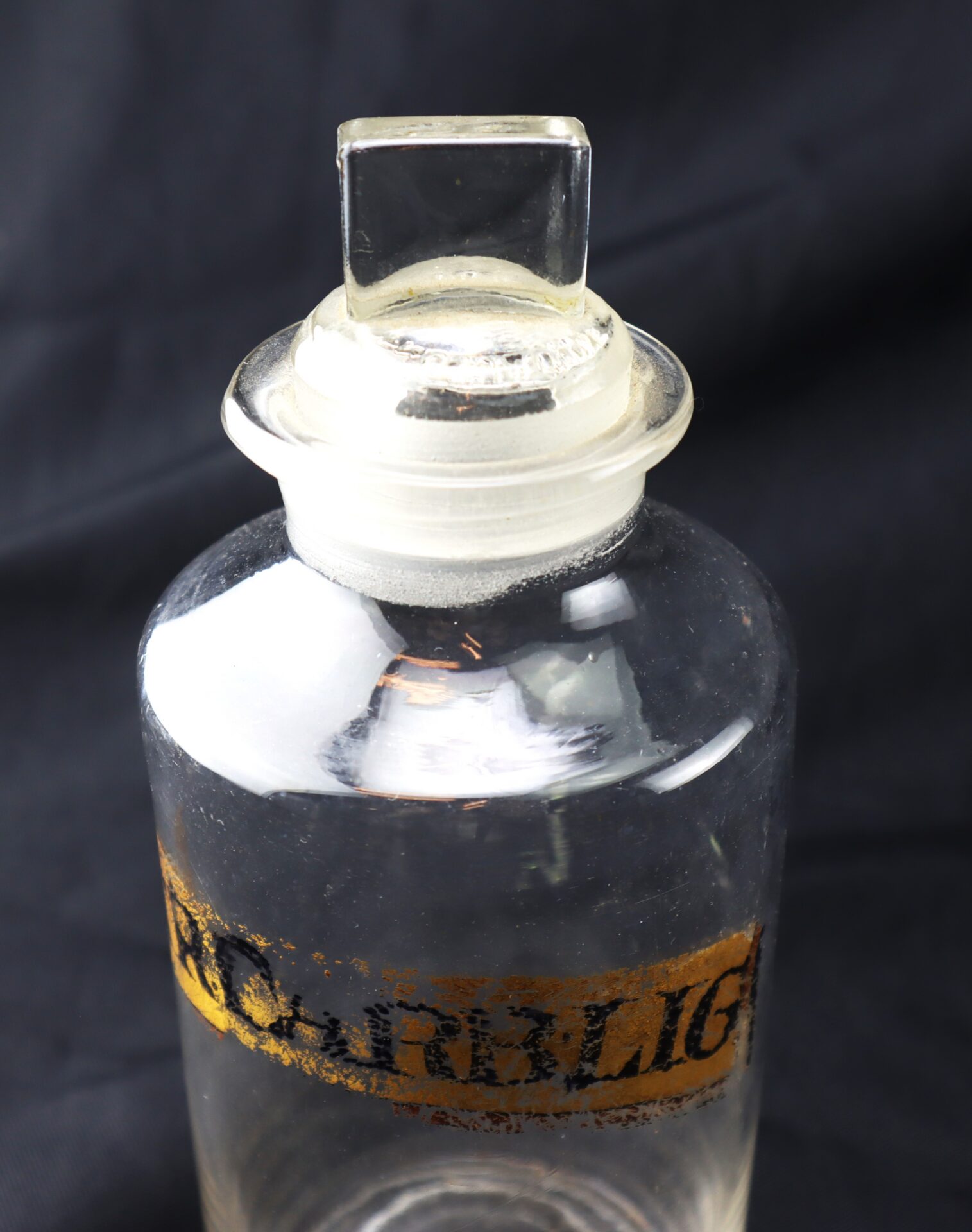 A clear glass bottle with a square glass stopper and a partially worn yellow label against a dark background.
