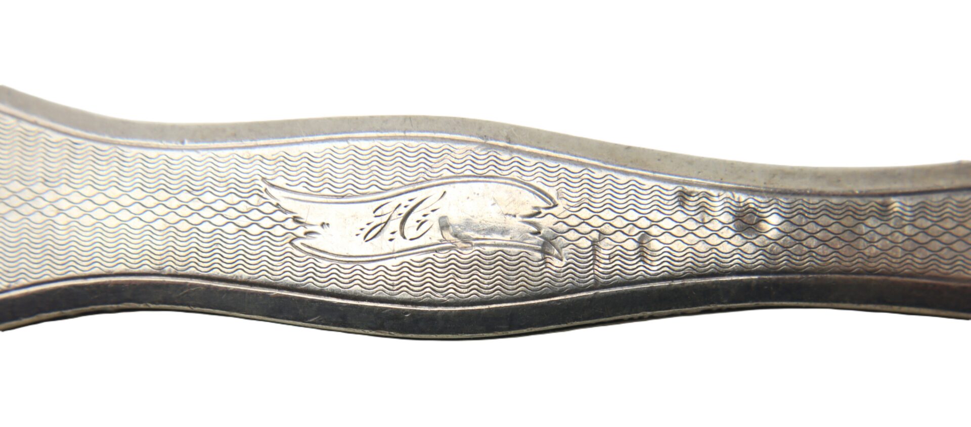 Close-up of a silver utensil handle with a wavy edge, textured pattern, and an engraved script letter “H” inside a decorative cartouche.