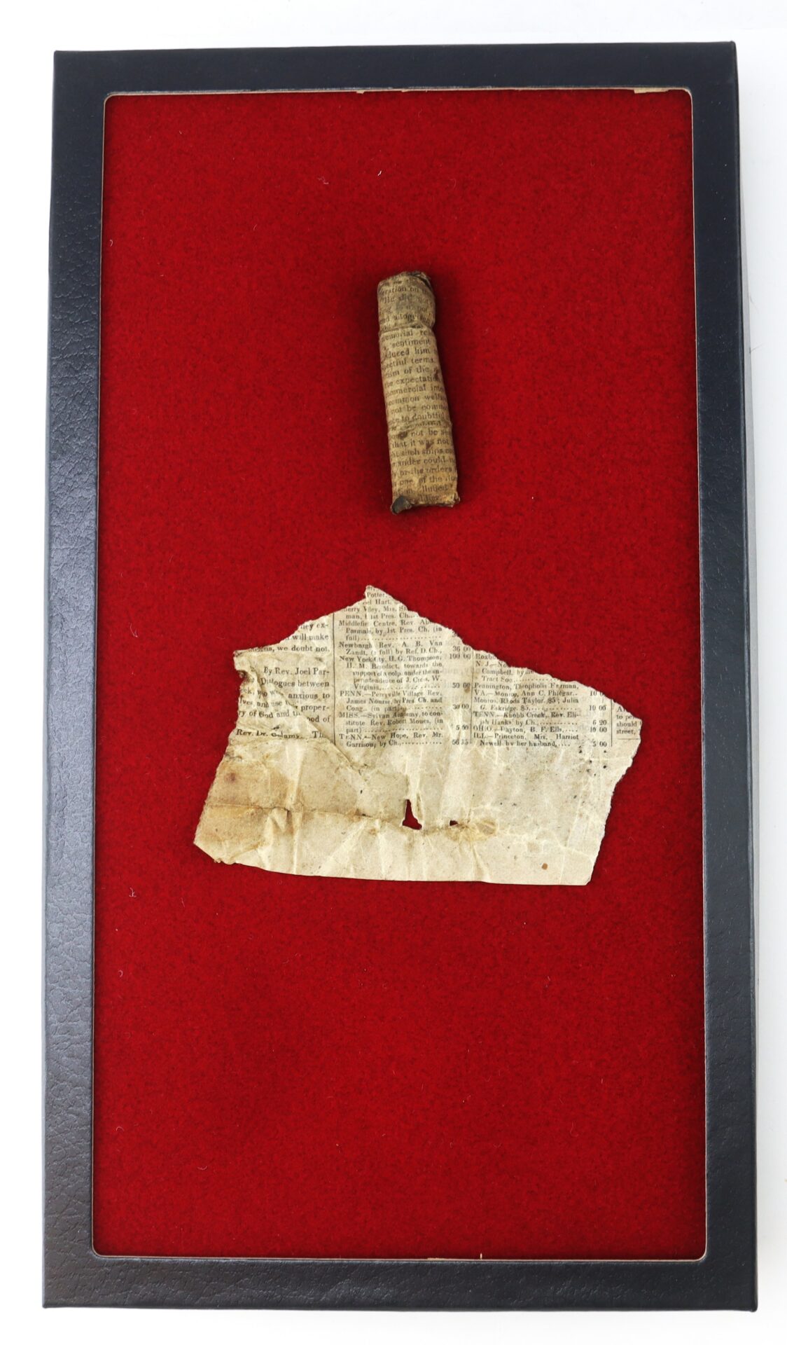 A small scroll and a torn sheet of old paper are displayed on a red velvet background in a black frame.