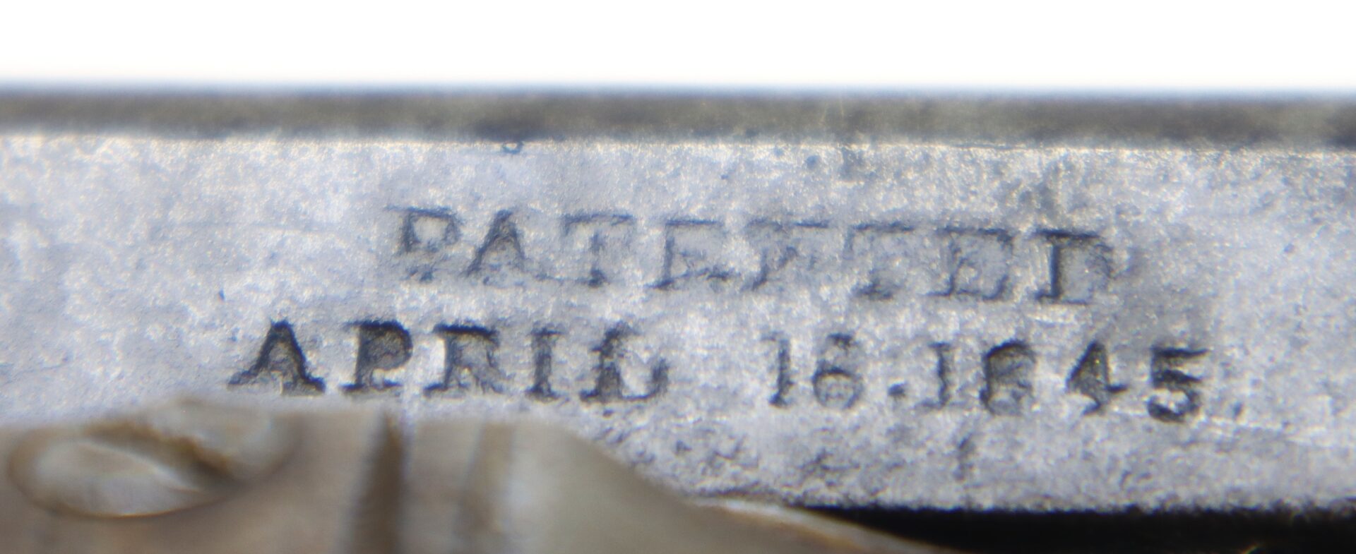 Close-up of metal surface engraved with the text “PATENTED APRIL 16.1845.” Some areas of the surface show wear and discoloration.